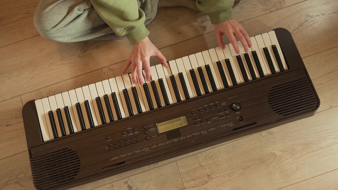 En scen från hög vinkel visar en person som sitter på träplankor, med fingrar som rör sig över kompakta elektroniska tangenter under avslappnad hemövning, med mjukt varmt ljus, texturerade kläder och en intim stämning.