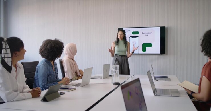 Female presenter in green pointing at screen showing app mockup sparking clapping in meeting room