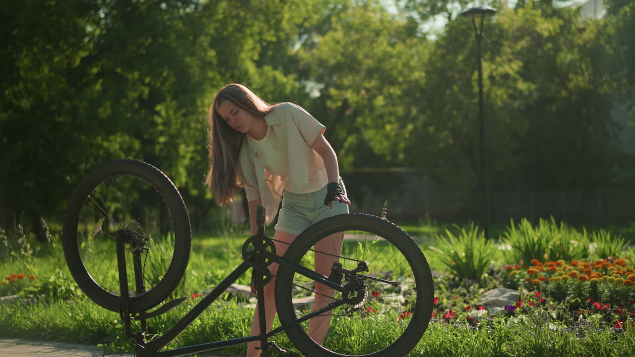 여성 자전거 타는 여성은 빛 공원 설정에서 활기찬 정원 에 거꾸로 자전거의 앞과 뒷 타이어를 조정합니다. 울창한 녹색 나무와 다채로운 꽃은 조용하고 흐릿한 배경을 제공합니다.
