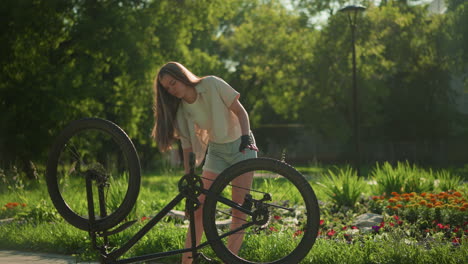 motociclista femenina ajusta las llantas delanteras y traseras de una bicicleta al revés al lado de un jardín vibrante en un entorno de parque soleado, árboles verdes exuberantes y flores coloridas proporcionan un fondo sereno y borroso
