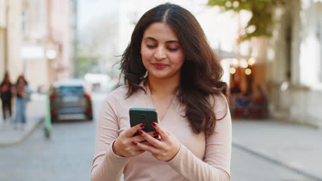 Indian-woman-use-smartphone-typing-text-messages-browsing-internet-finishing-work-looking-at-camera