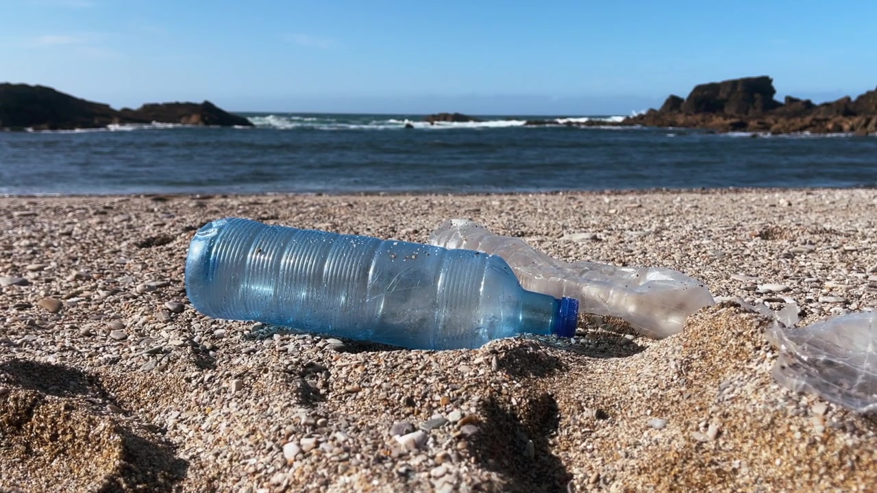 Sininen muovipullo ja muoviroskaa saastuttamassa rantaa, valtameren aallot taustalla.