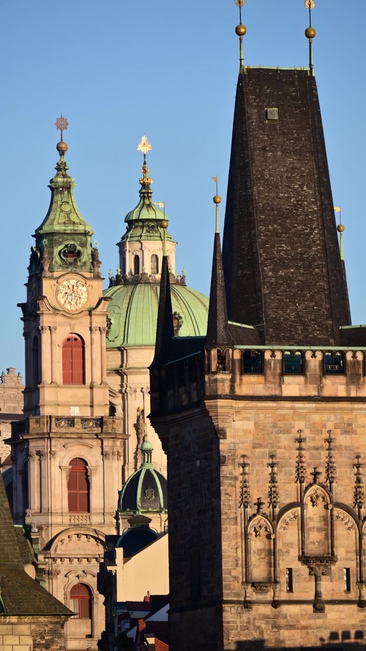 Primo piano verticale della torre dell'orologio e della cupola verde della Chiesa di San Nicola accanto a una scura guglia gotica, Praga, Repubblica Ceca