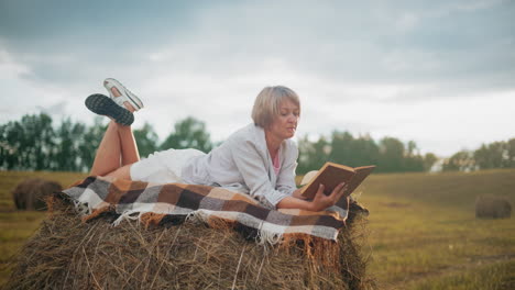 woman resting on hay bale in open field, reading a book while wrapped in a checkered blanket, enjoying peaceful moment surrounded by nature in rural countryside setting