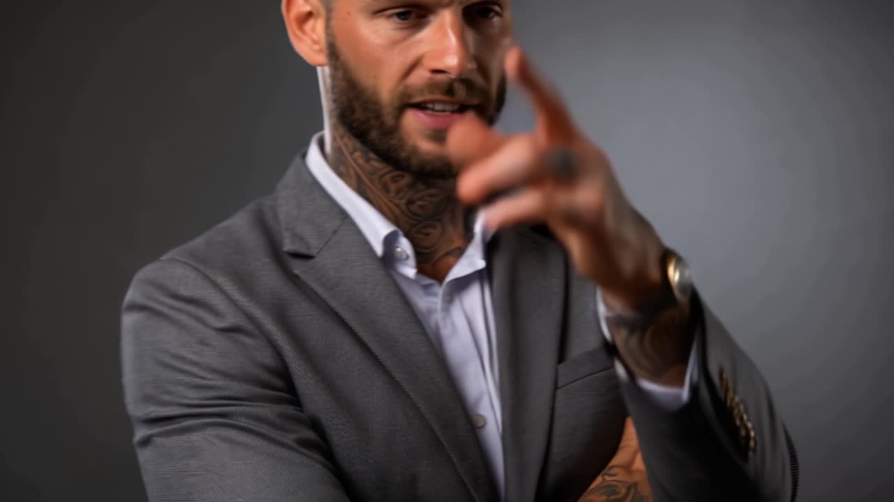 Homem com tatuagens veste terno, braços cruzados. Ele gesticula enquanto fala, mostrando confiança. Sua expressão sugere autoridade e foco. Ótimo para negócios, liderança ou temas motivacionais.