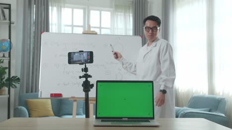 asian male teacher with glasses and green screen laptop shoots video by smartphone camera while teaching chemist at home