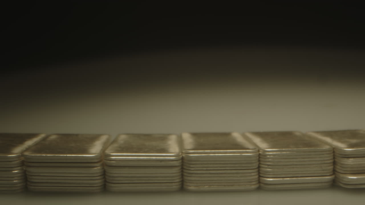 Dolly in to multiple stacks of silver bars illuminated by a spotlight in a dark room. The precious metal plates come into focus as the camera moves closer