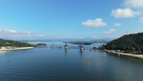 Guaratuba Bay in Paraná, Brazil, capturing the construction of the Ponte de Guaratuba bridge linking Guaratuba and Matinhos, with coastal hills and calm waters below, high drone shot