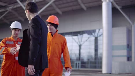 Manager-in-a-suit-giving-envelopes-with-money-to-workers-in-orange-uniform-and-helmets.-Successful-finish-of-the-project.-Shot-in-4K