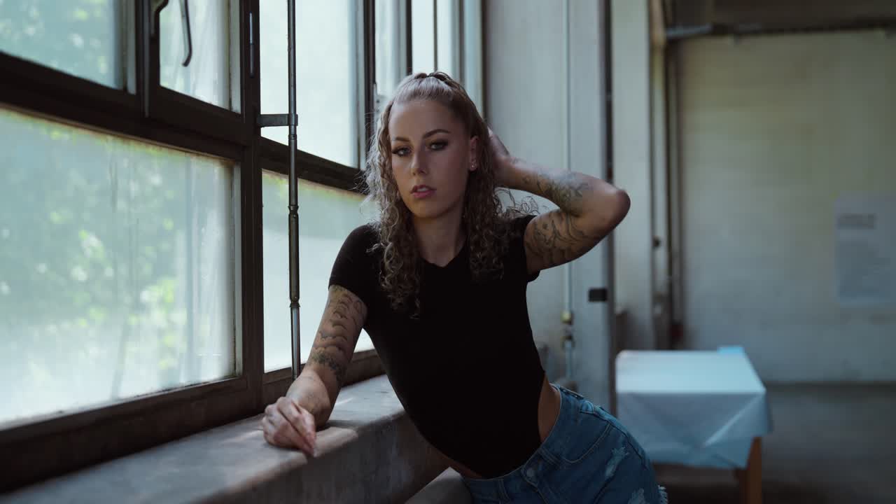 Tattooed woman posing confidently by industrial windows in natural light