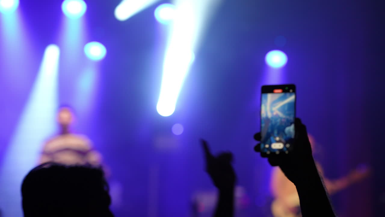 Audience member holding smartphone to capture performance on stage, illuminated by colorful stage lights, creating an energetic atmosphere at a live music event