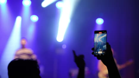 audience member holding smartphone to capture performance on stage, illuminated by colorful stage lights, creating an energetic atmosphere at a live music event
