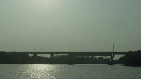 Río-Iluminado-Por-El-Sol-Brillando-Bajo-La-Silueta-De-Un-Gran-Puente,-Barcos-Amarrados-Cerca-De-Una-Exuberante-Vegetación