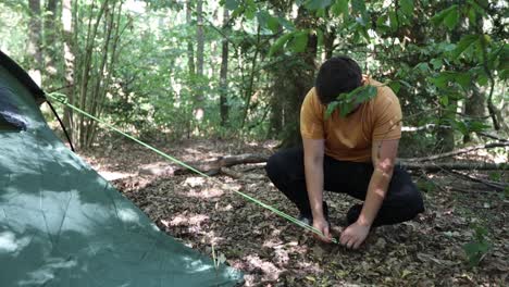un hombre decepcionado monta una tienda de campaña en un campamento en el bosque pero no logra poner el gancho en el suelo dos veces y deja caer la cuerda
