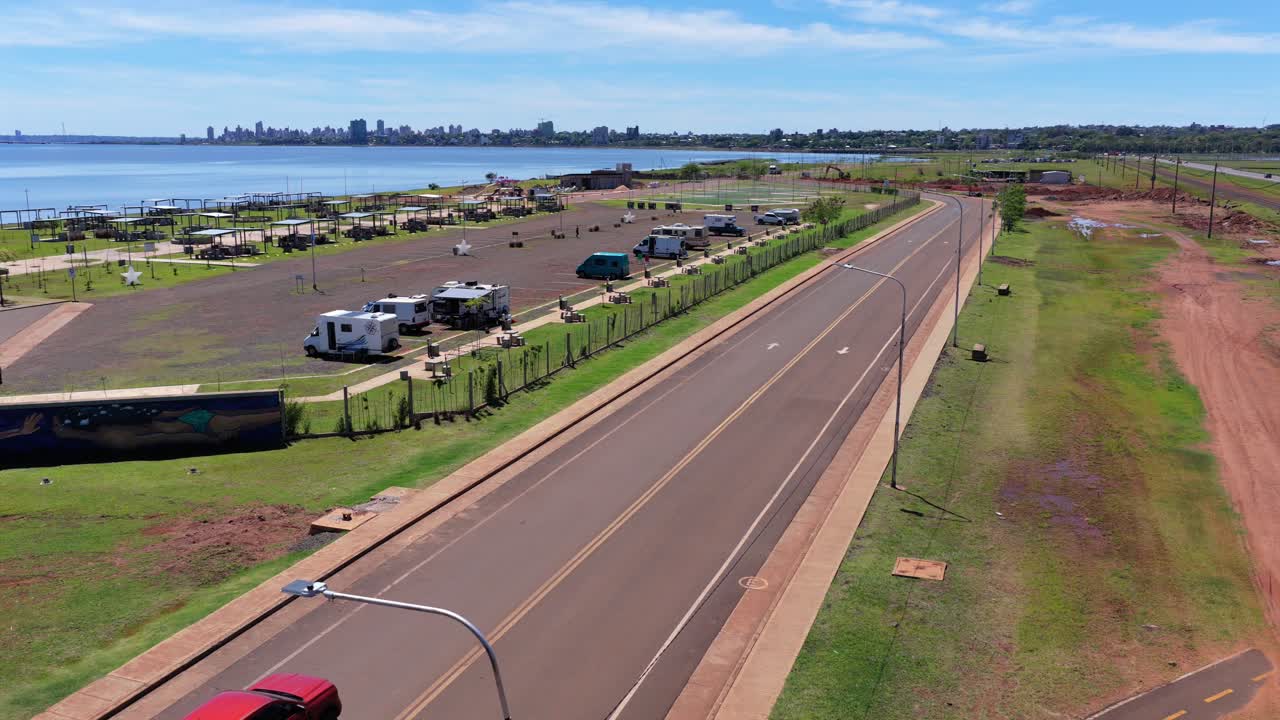 Descending drone closes in on red pickup truck driving next to RV park with water and city views. Misiones, Argentina