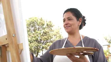 Happy-biracial-senior-woman-painting-picture-and-smiling-in-sunny-garden,-slow-motion,-copy-space