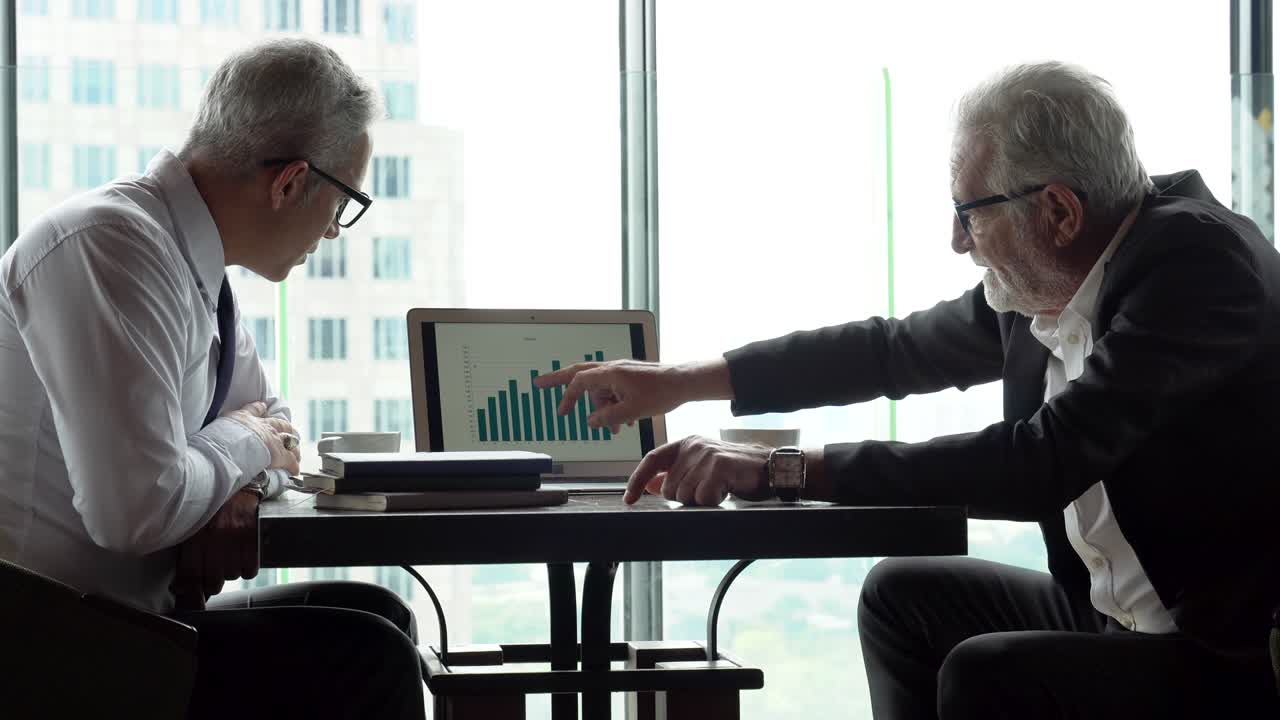 dos hombres de negocios sénior en la reunión apuntando gráfico en el portátil analizando la información de datos. anciano ceo discutiendo con el cliente en el café. serio gerente maduro hablando por la ventana sobre el paisaje urbano de lujo
