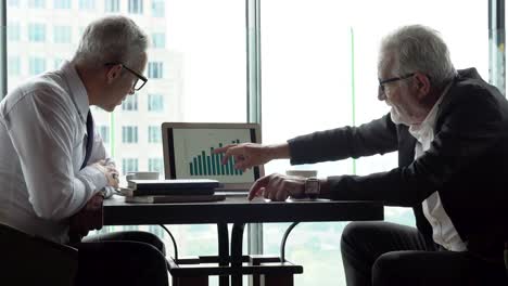two senior businessman in meeting pointing graph in laptop analyzing  data information . old man ceo discussing with client in cafe. serious mature manager talking by window over cityscape luxury