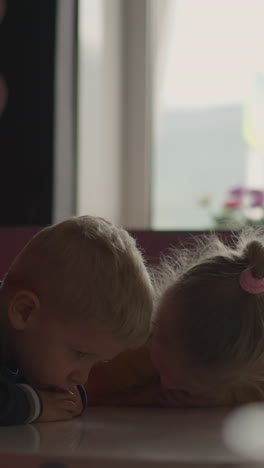 offended little brother and sister bend putting heads on empty table sitting together in restaurant hallway against window and board panel closeup