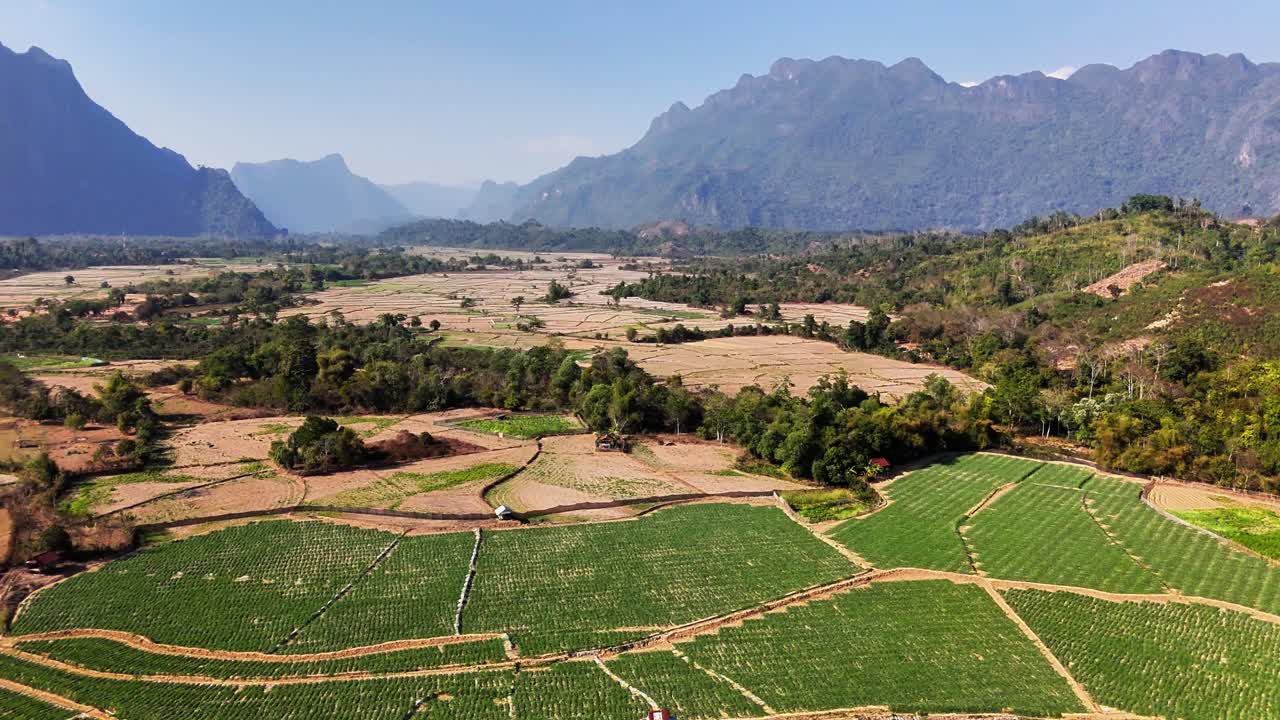 Il drone scivola sopra il villaggio di Pha Tang lungo il fiume Nam Song a Vang Vieng, Laos, con campi terrazzati, case tradizionali con tetto laotiano e la cresta calcarea Pha Ngern che si erge sopra la valle.