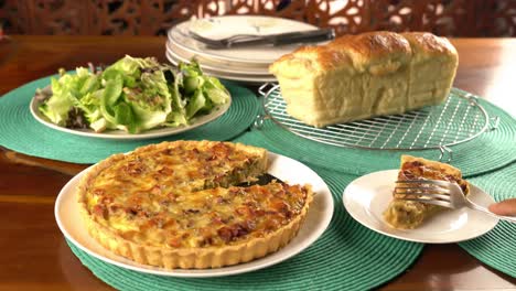 Using a metal fork taking a bite of green pea and bacon quiche in white ceramic plate on blue table mat.