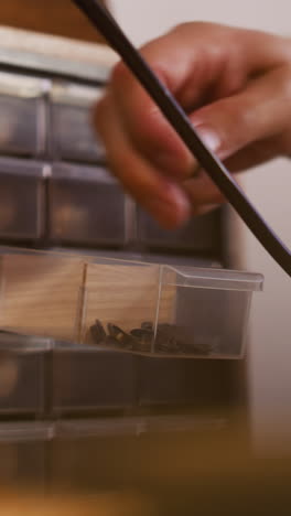 man takes metal rivet from small plastic drawer in workshop closeup. master chooses fastening for craft at workplace. manufacture of accessories