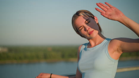 young lady resting right hand on iron railing, using left hand to block sunlight from eyes, squinting under bright sun, background blurred with distant greenery