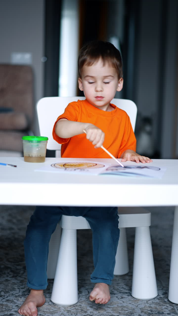 Concentrated Caucasian baby boy painting on a piece of paper. Toddler doing art at home. Vertical video.