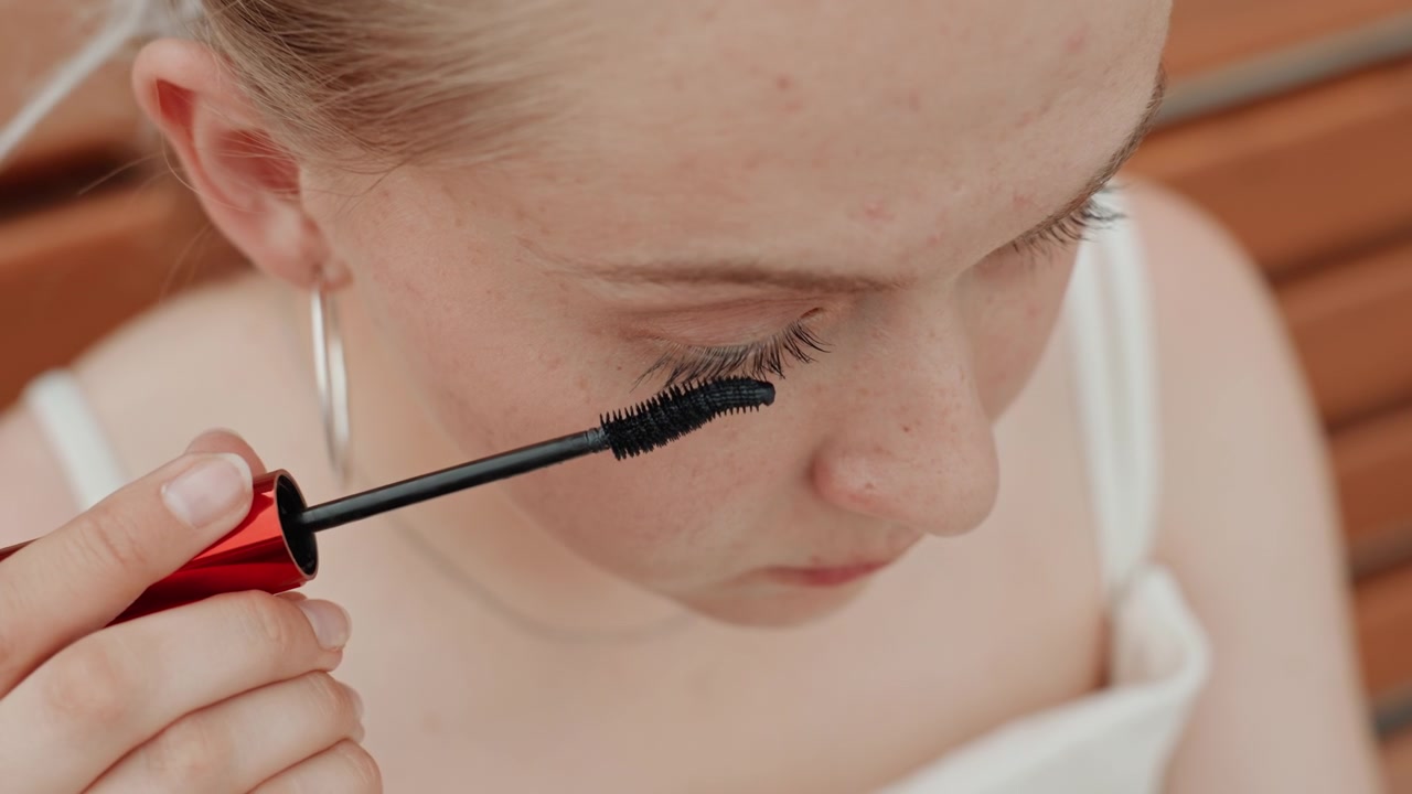 Young woman applies black mascara to her eyelashes in close up view focusing on precise movement of applying makeup. Woman wears hoop earrings and white top with wooden slats visible