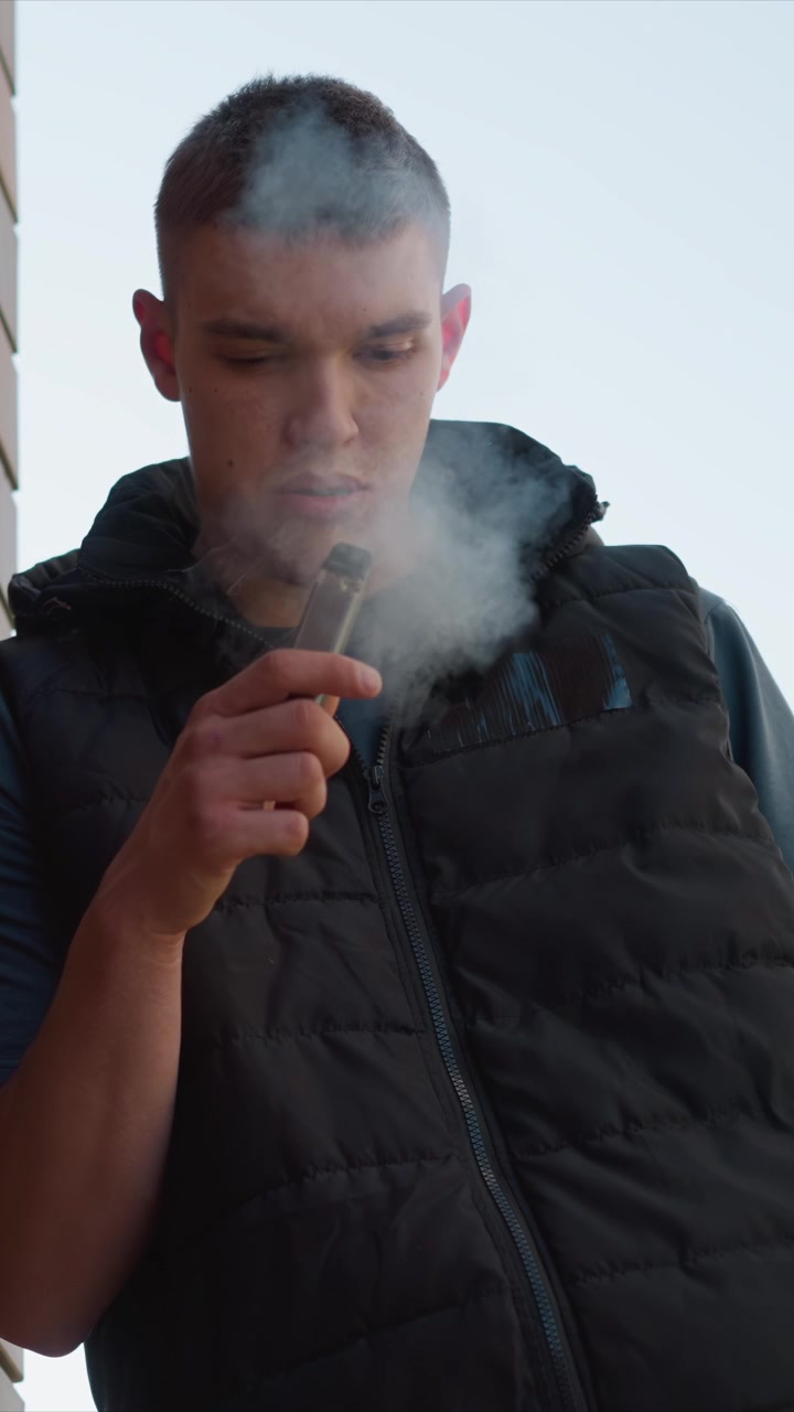 Primo piano di un uomo che tiene e usa un dispositivo da svapo esalando vapore. L'uomo indossa un gilet imbottito nero sopra una camicia blu in un ambiente esterno con uno sfondo di cielo sereno. L'azione dello svapo è il fulcro centrale.