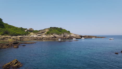 Zoom-In-Shot-Of-Blue-Sea-With-Rocky-Coast-While-Some-Yachts-Are-Anchored-Near-The-Shore-And-People-Swimming