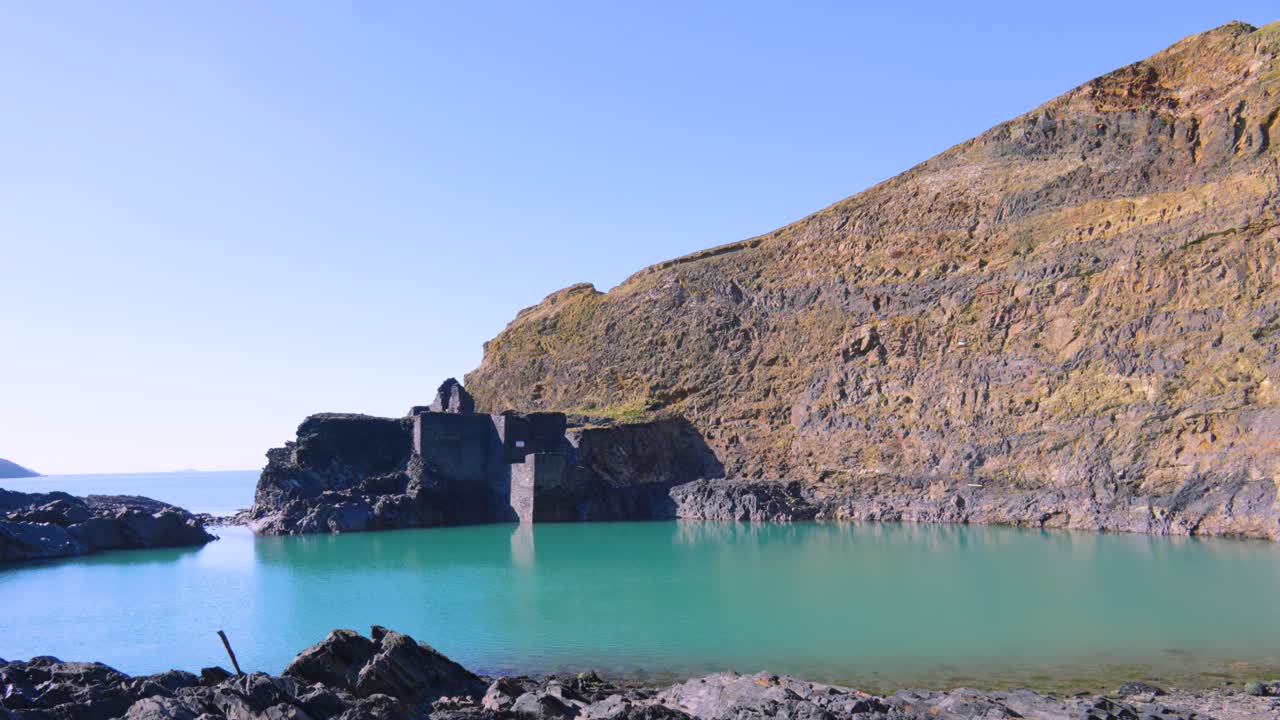 Blue Lagoon in Old Quarry with Tidal Sea Water on Bright Hot Sunny Day with Rocks and Industrial Ruins Next to Sheer Coastal Cliffs and Ocean in Background 4K.