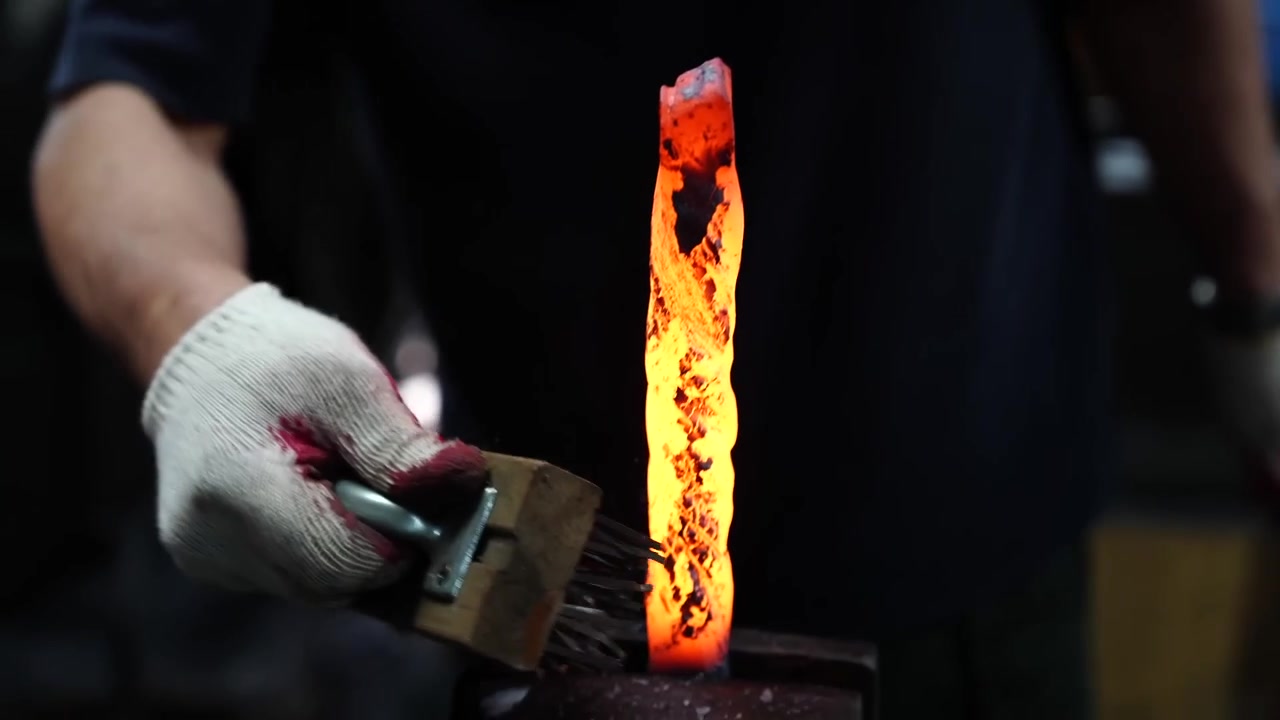 Craftsman brushing scale off a vertical glowing steel bar with a wire brush, sending sparks across the anvil