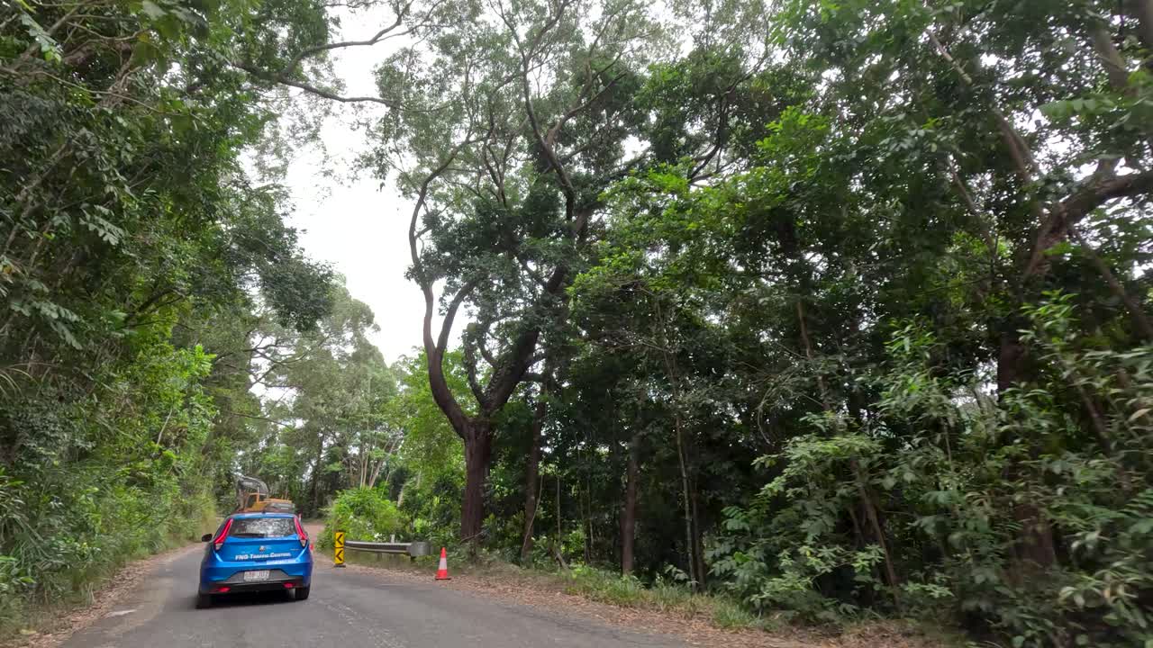 Blue car travels winding rainforest road, lush greenery, overcast daylight, smooth forward camera movement