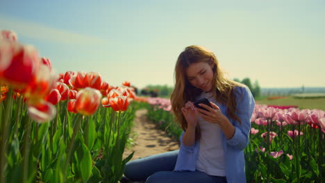 Happy-woman-touching-screen-of-mobile-phone-while-sitting-on-road-at-tulip-field