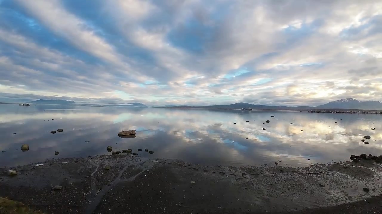 Narrow tidal creek winding through grassy shoreline toward the mirror-still Última Esperanza Fjord, with distant mountains and a pier visible near Puerto Natales, Chile, FPV aerial