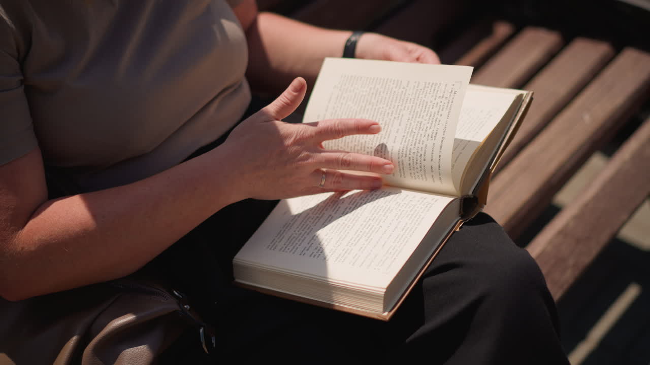 Close up of student seated outdoors on wooden bench flipping to new page in open book under sunlight calm mood relaxed summer atmosphere showing focus learning curiosity