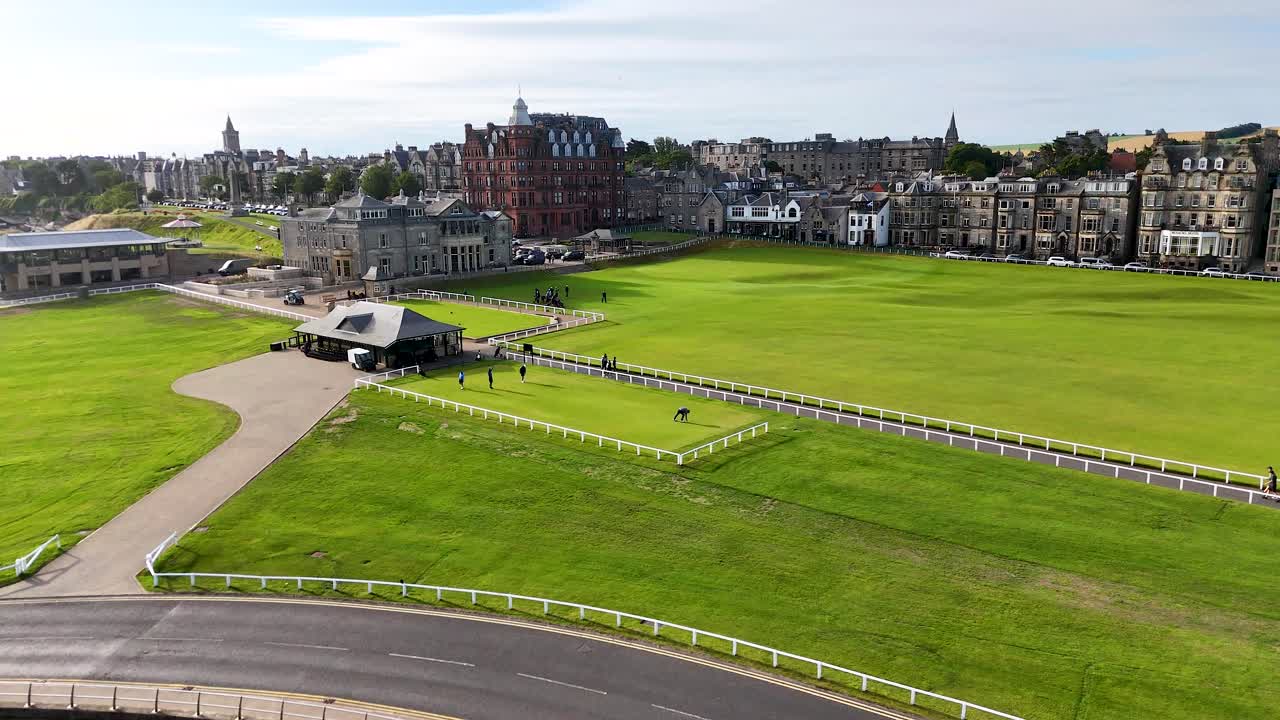 Aerial tracking shot of a blue car driving along a curved road beside the Old Course, with Victorian architecture and green lawns under daylight