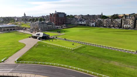 aerial tracking shot of a blue car driving along a curved road beside the old course, with victorian architecture and green lawns under daylight