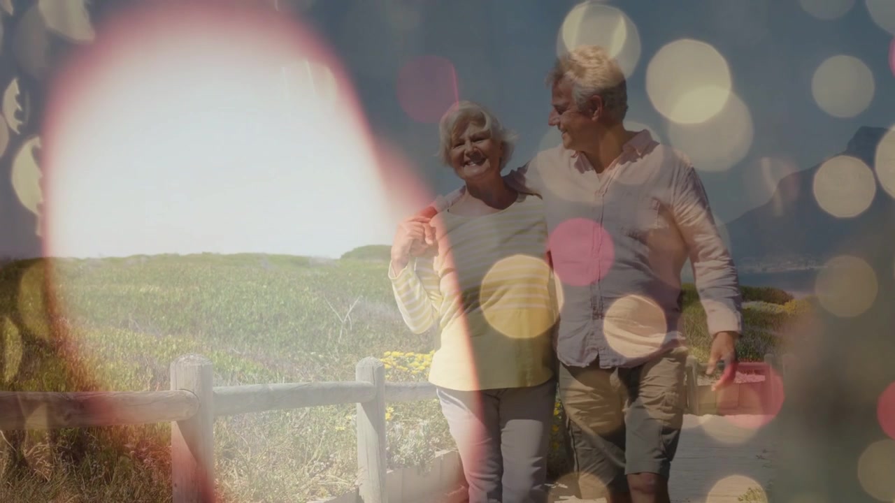 Pareja mayor caminando por un sendero costero, el hombre sujetando el brazo de su esposa, con un efecto de destello que ilumina el bokeh, por su salud.