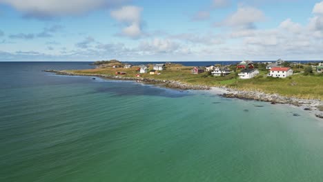 Flying-above-paradise-beach-with-bright-white-sand-and-waves-on-turquoise-water-in-Lofoten-islands-in-Northern-Norway-revealing-the-scenery-of-typical-red-houses-on-a-sunny-summer-day