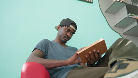 Dark skin boy in cap sits on red couch with crossed leg, holding open book, reading calmly in bright indoor room with turquoise wall and bookshelf, sneaker in foreground