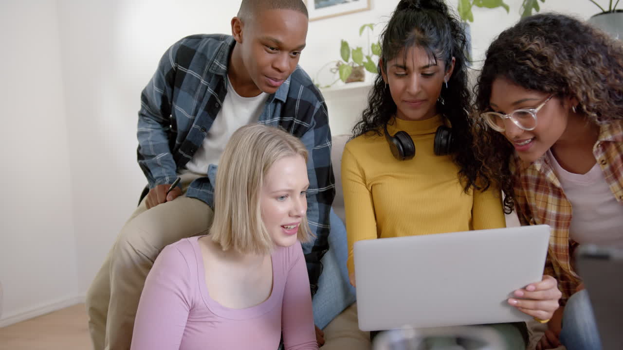Happy diverse group of teenage friends sitting on couch and using laptop at home, slow motion
