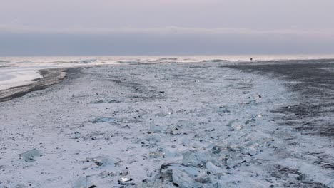 Aerial-view-over-ocean-waves-crashing-on-diamond-beach,-covered-in-snow,-at-sunset