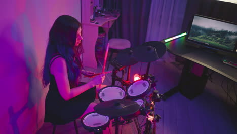 young girl seated at electronic drum kit in studio lit with purple glow, playing with drumsticks while looking dull, monitor screen and desk setup visible nearby with musical equipment