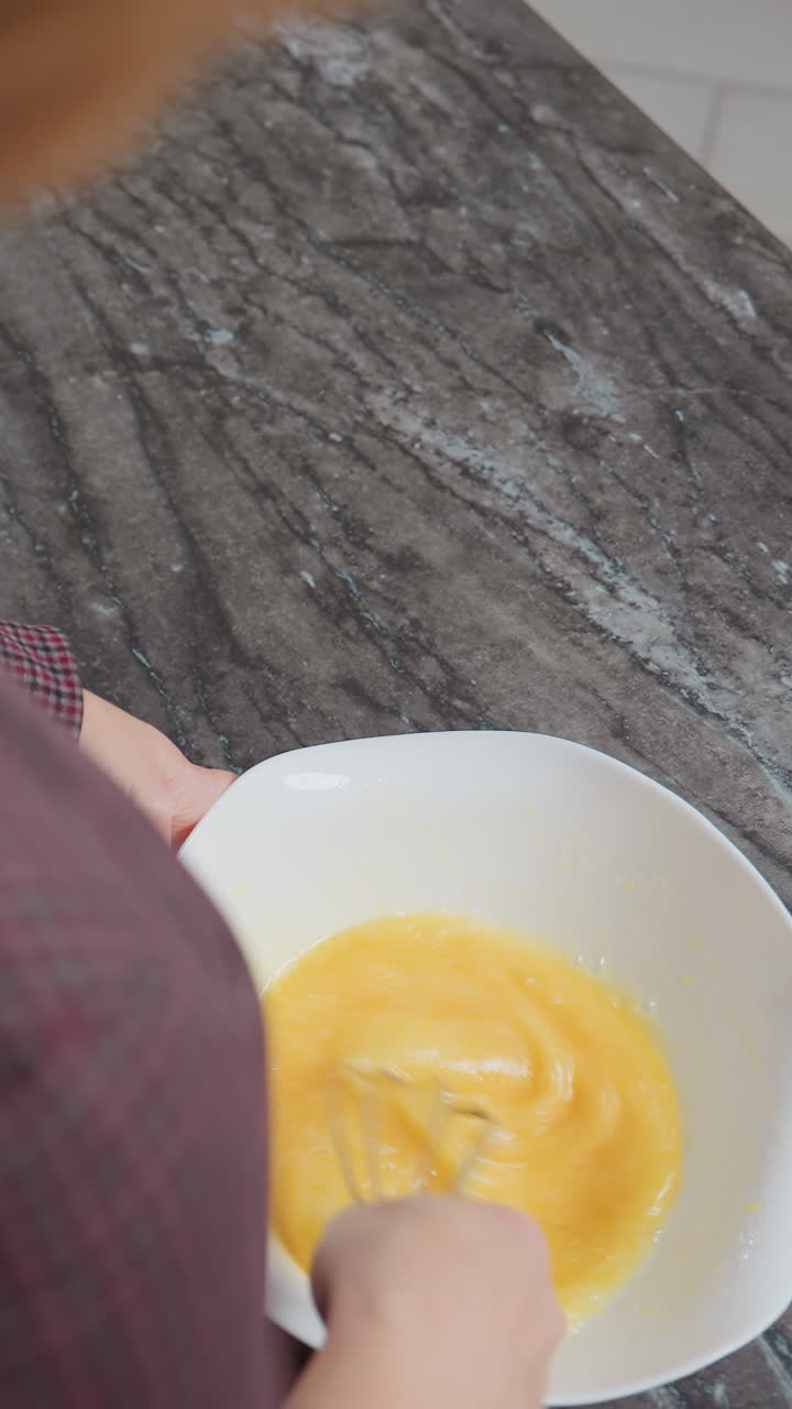 primer plano de una mujer de cabello dorado con vestido a cuadros moviendo un huevo en un cuenco, levanta brevemente el batidor antes de continuar mezclando, el fondo presenta una encimera de mármol