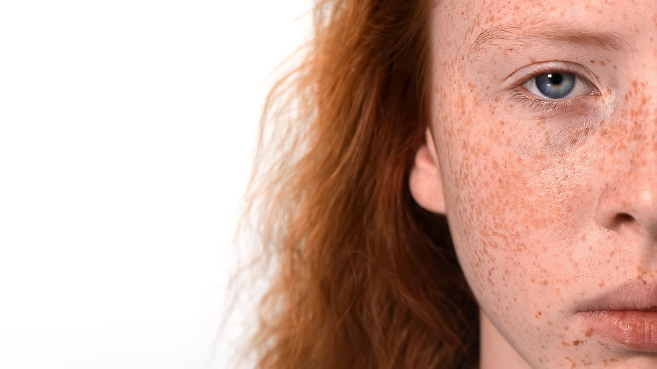 Red haired girl with lots of freckles on the face open the blue eye and looking at the camera. White background. Place for text. Close up