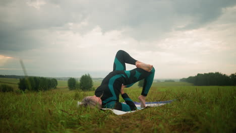 mujer de traje negro verde acostada en una alfombra de yoga practicando la postura del puente con las manos apoyando su cintura, sentada en un vasto campo de hierba bajo un cielo nublado, con árboles en la distancia