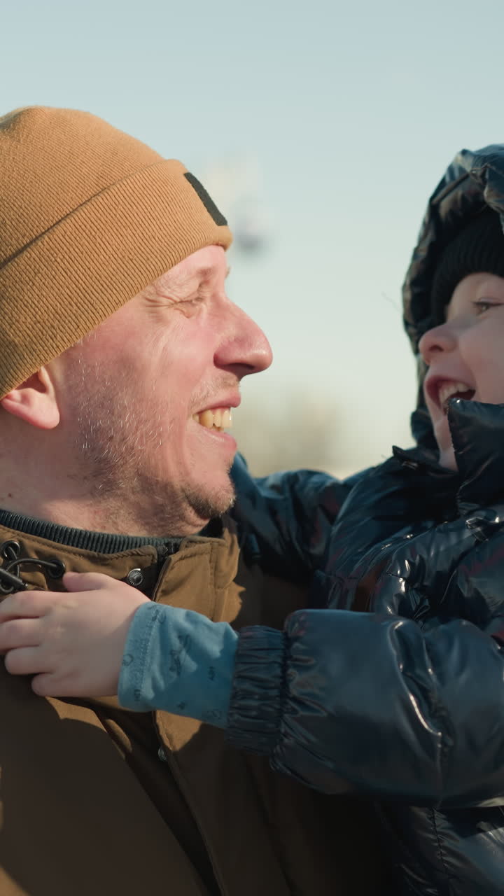 отец носит своего сына, а ребенок игриво привлекает его внимание, создавая момент радостного взаимодействия, отец носит коричневую куртку, а сын одет в черную пушистую куртку