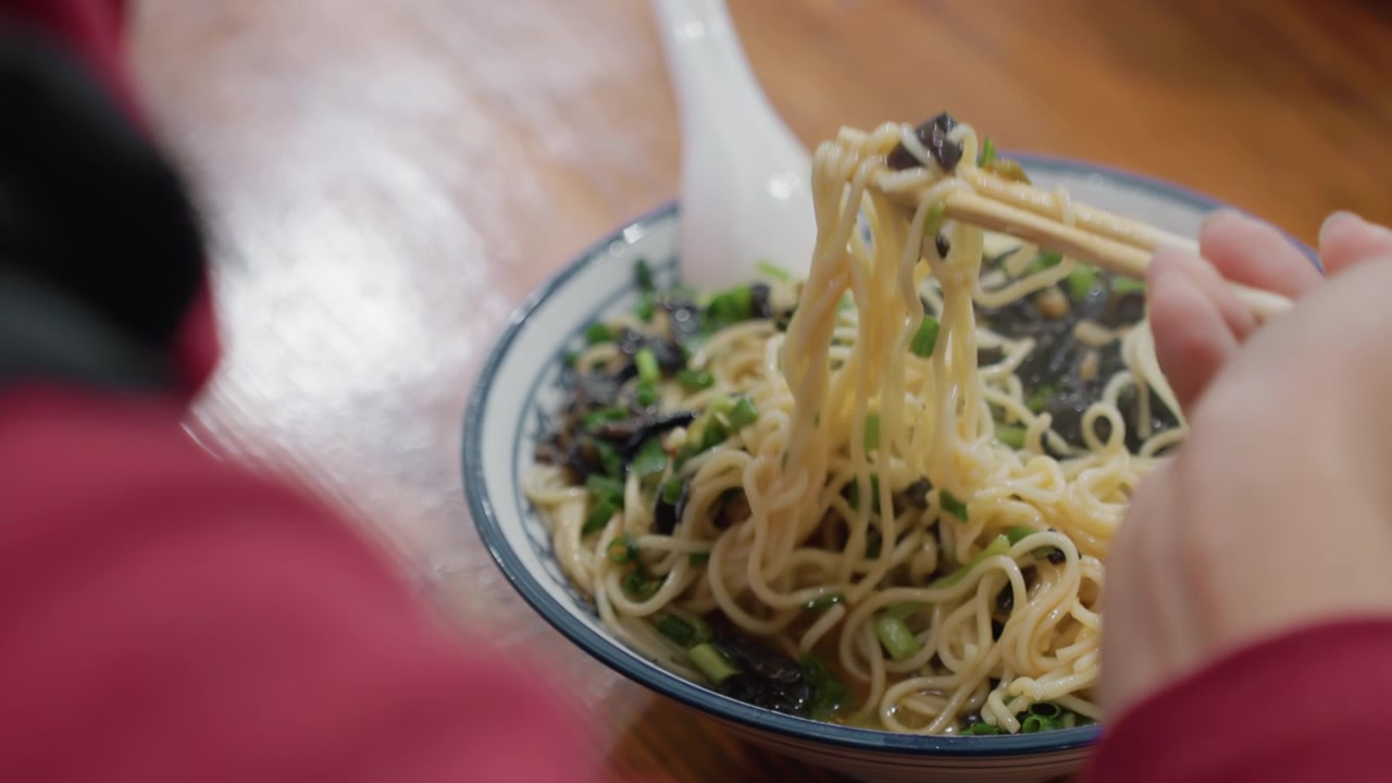 Tangan Close-up Memutar Mangkuk Mi, Sumpit Mengangkat Helai Mi di Atas Mangkuk Keramik dengan Hiasan Daun Bawang dan Jamur, Meja Kayu, Lengan Jaket Merah, Momen Makan Nyaman yang Alami dengan Tekstur dan Uap